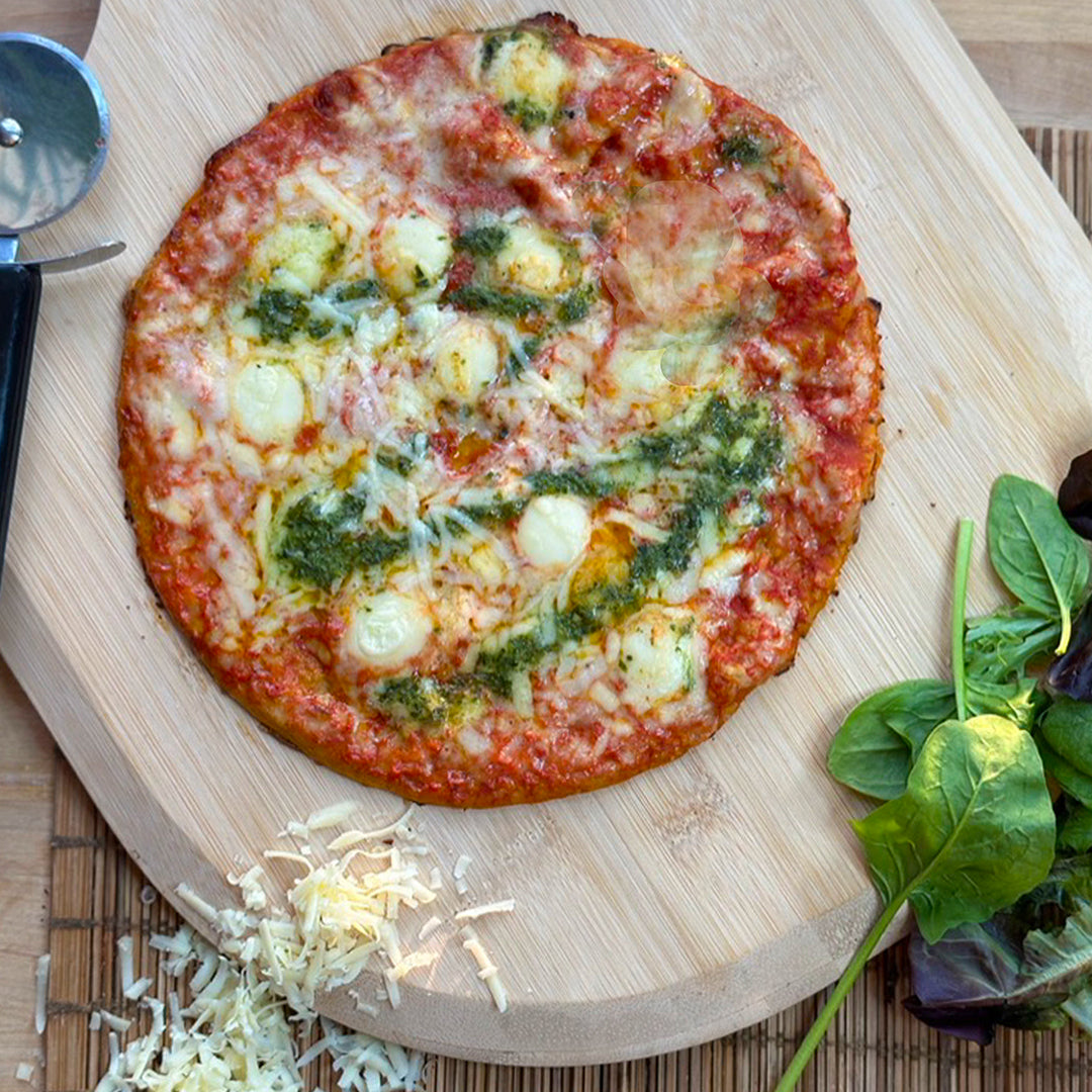 Picture - Cooked low carb keto cheese and pesto pizza fresh out the oven on a wooden chopping board. Pearls of mozzarella melting on top, drizzles with pesto. 